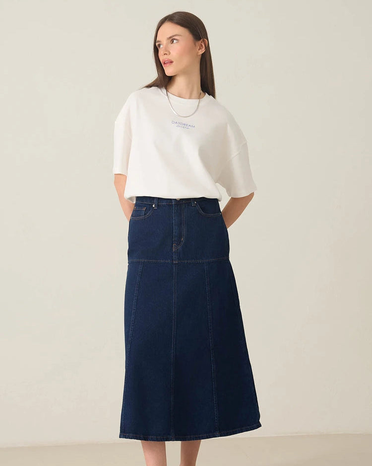 A woman modeling a white short-sleeve t-shirt and a dark navy blue denim midi skirt, paired with white sneakers, against a plain off-white studio background.